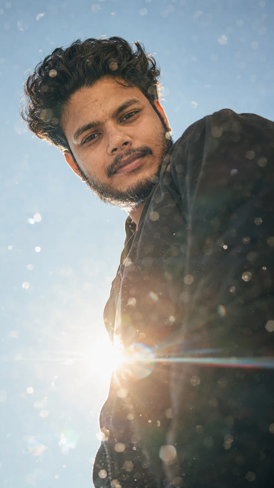 A low-angle portrait photograph capturing a young South Asian man with curly hair and a beard, looking down directly at the camera.