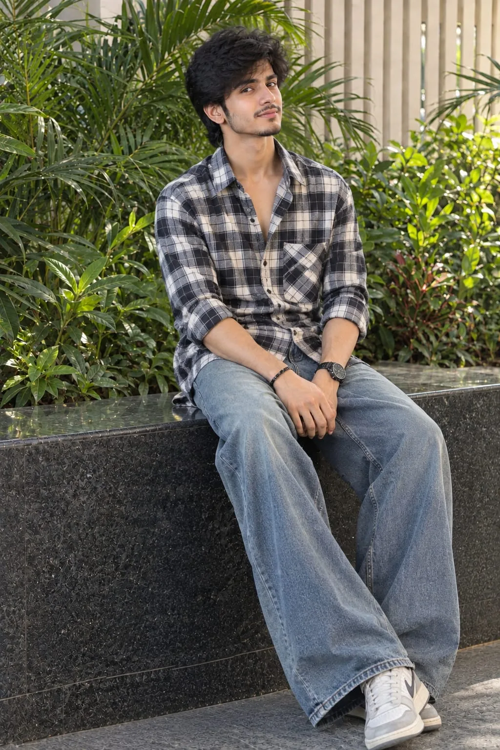 Ultra-realistic young South Asian man seated casually on a dark stone ledge, wearing an oversized black-and-white plaid shirt, loose