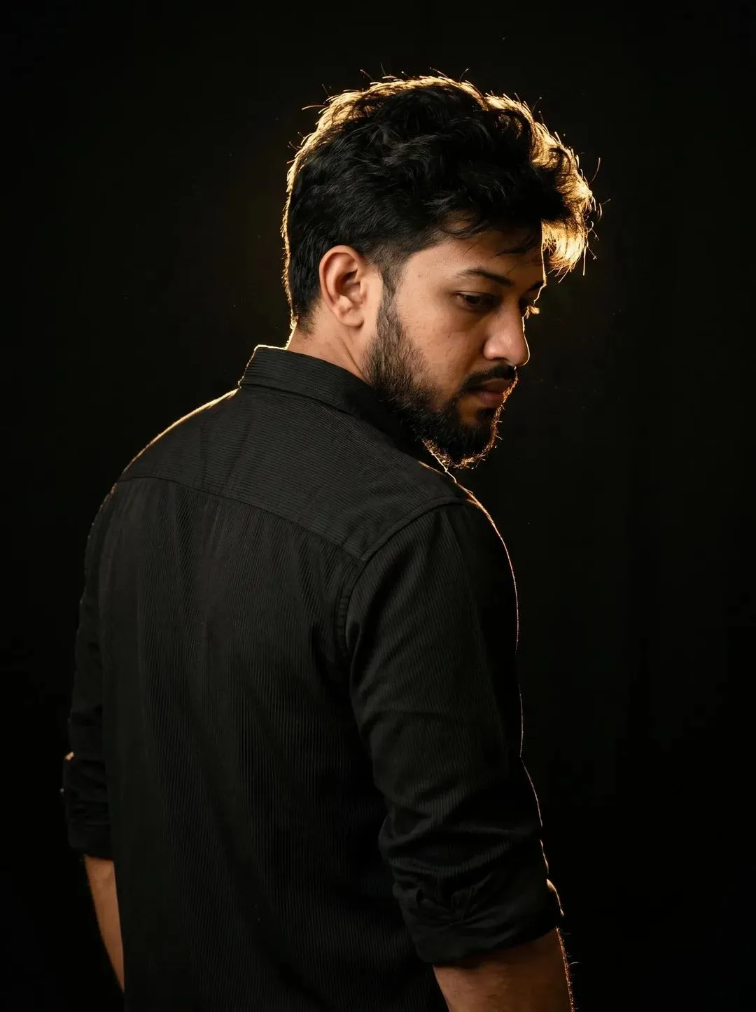 Create a cinematic ultra-realistic portrait of a young man standing with his back slightly turned toward the camera in a dark black studio