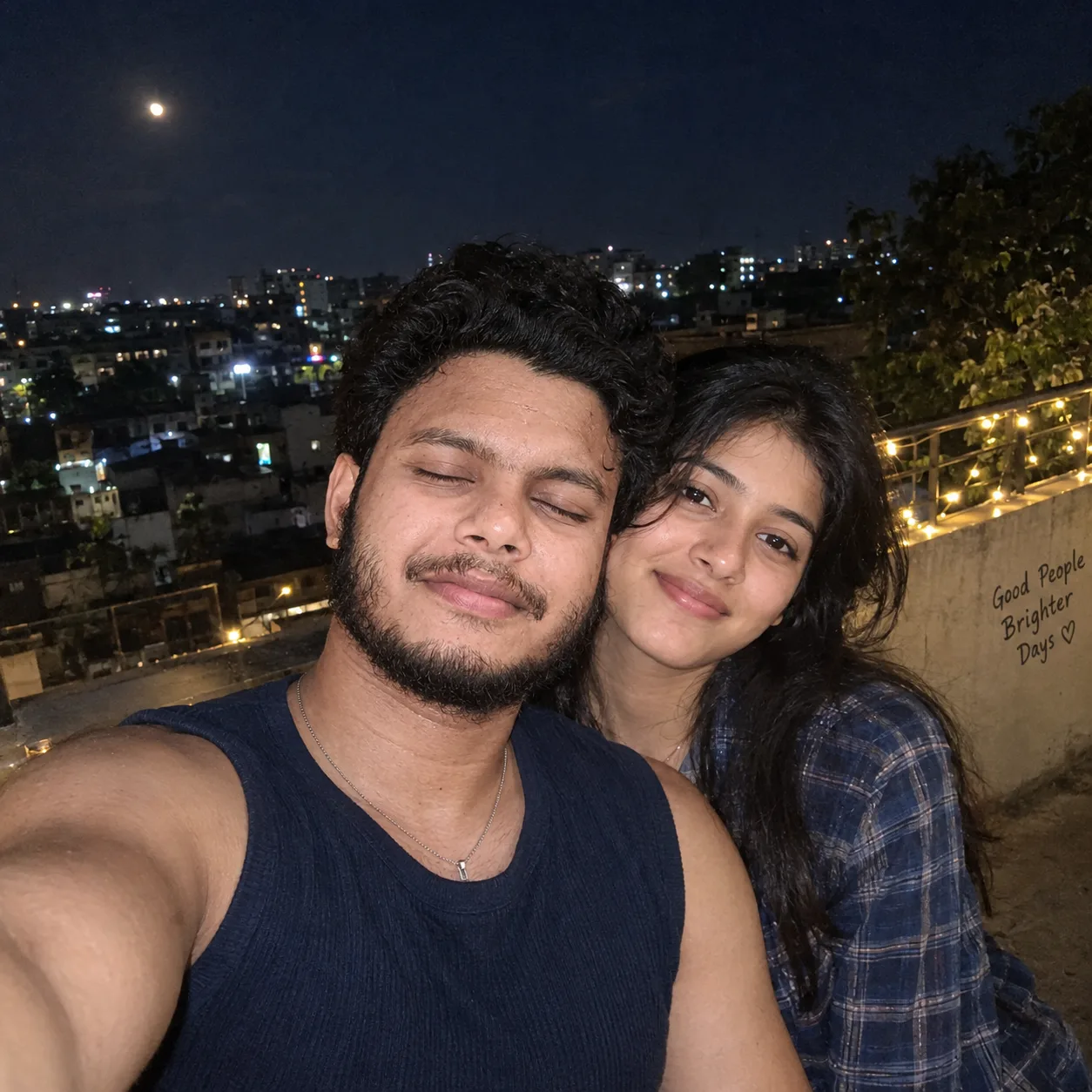 A young South Asian couple taking a close-up selfie on a rooftop at night.