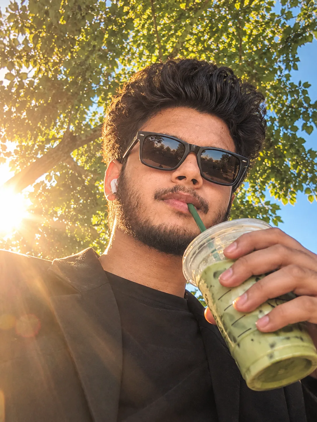 Prompt Low-angle selfie photo of a young man wearing dark square sunglasses and a wireless earbud, sipping an iced matcha latte through a