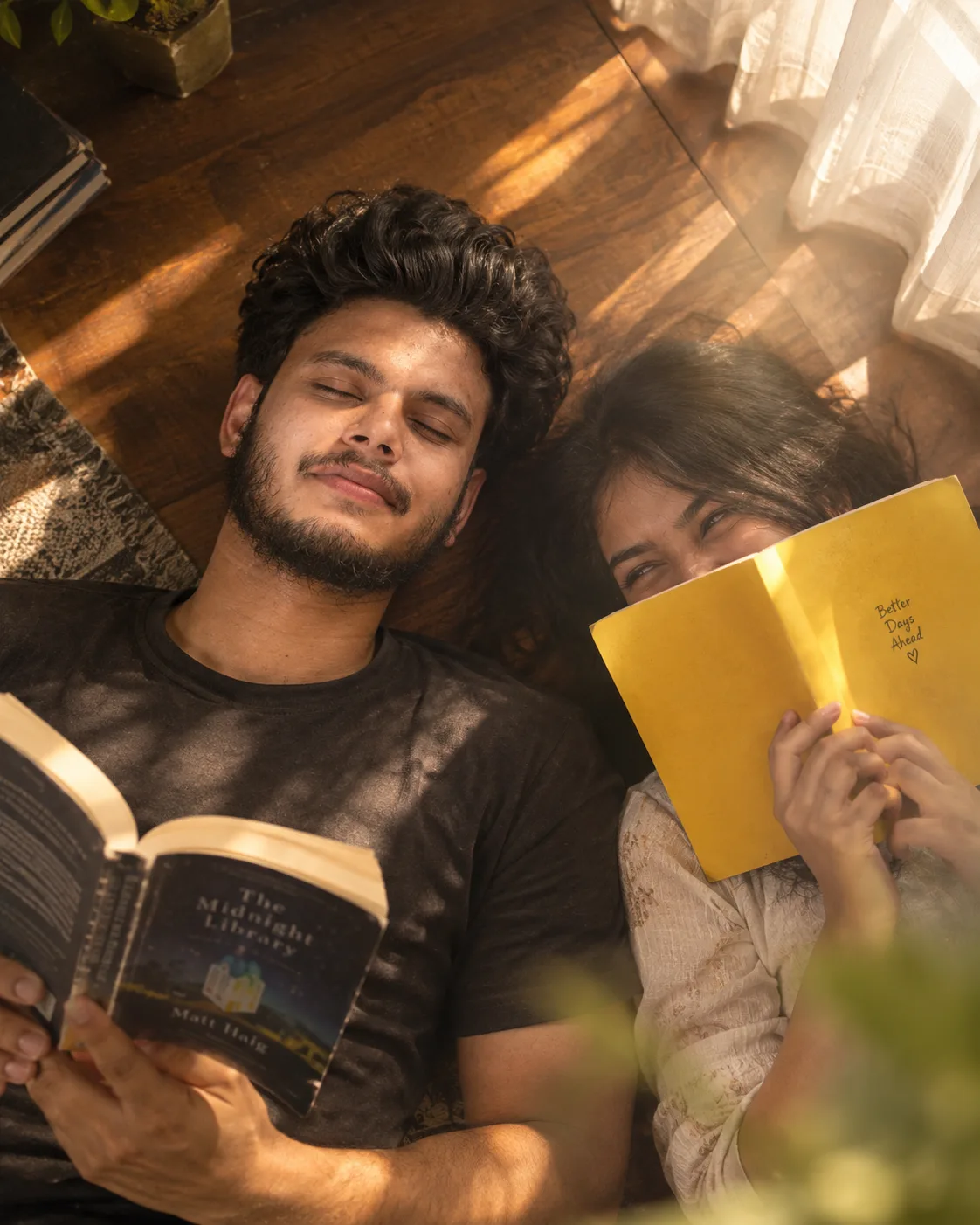 TOP-DOWN CANDID PORTRAIT, CURLY-HAIRED STUBBLED MAN RECLINES EYES-CLOSED WITH AN OPEN BOOK, WOMAN BESIDE HIM HIDES A LAUGH BEHIND A BRIGHT