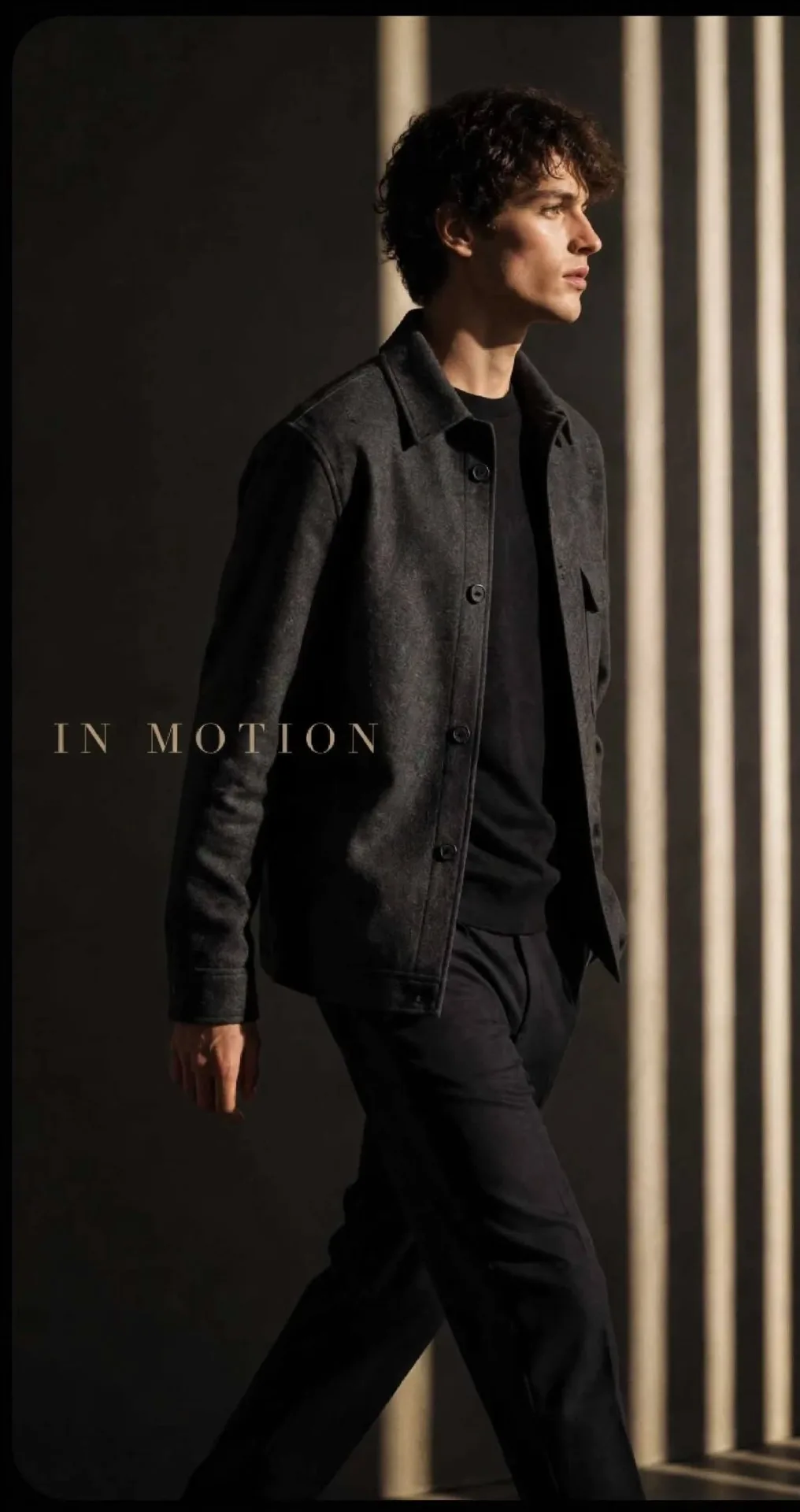 Cinematic fashion editorial portrait of a young man with curly dark hair, wearing a dark charcoal overshirt over black t-shirt and black