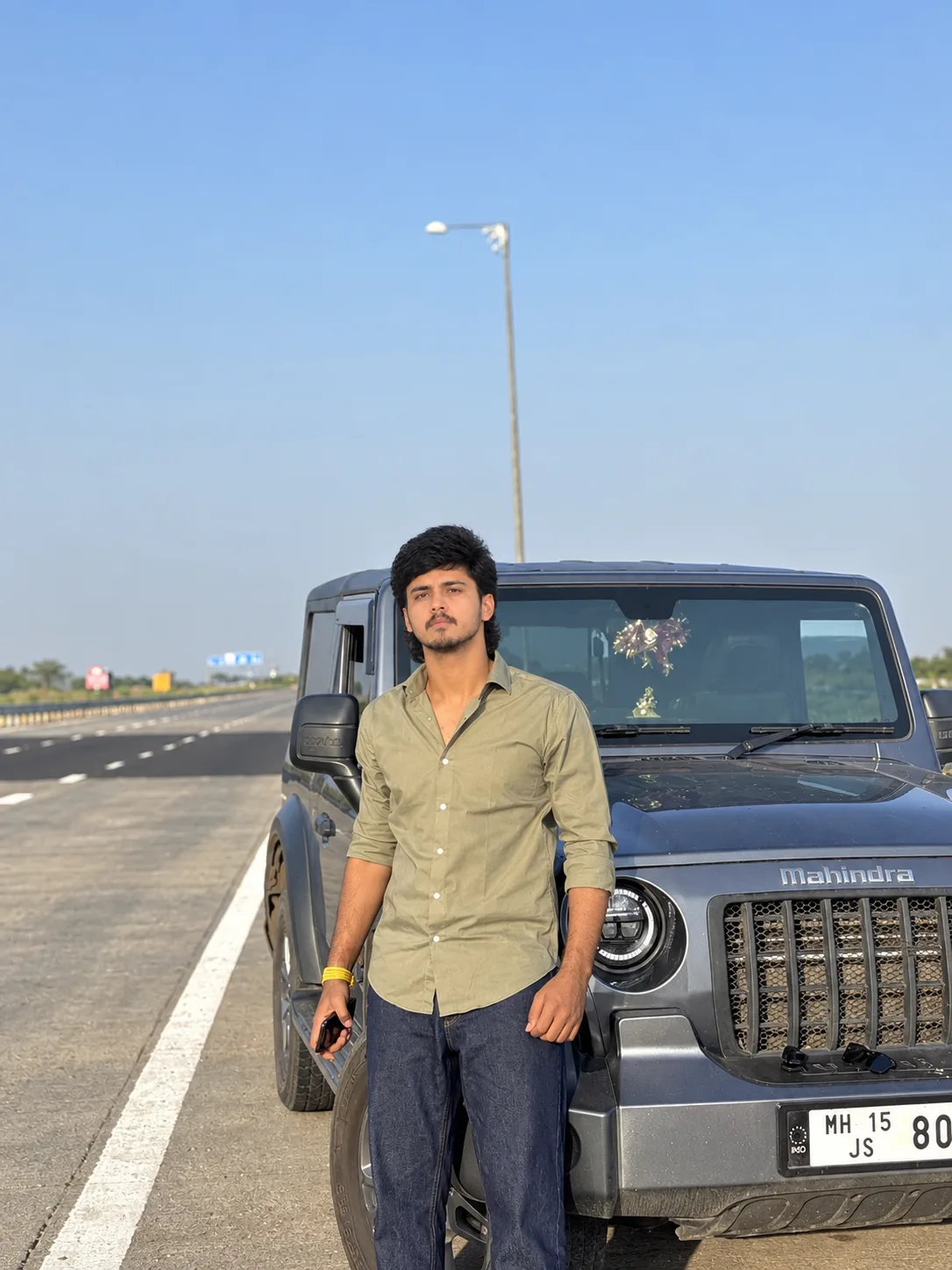 A photorealistic vertical portrait of a young South Asian man standing confidently on a wide open highway beside a dark charcoal-gray
