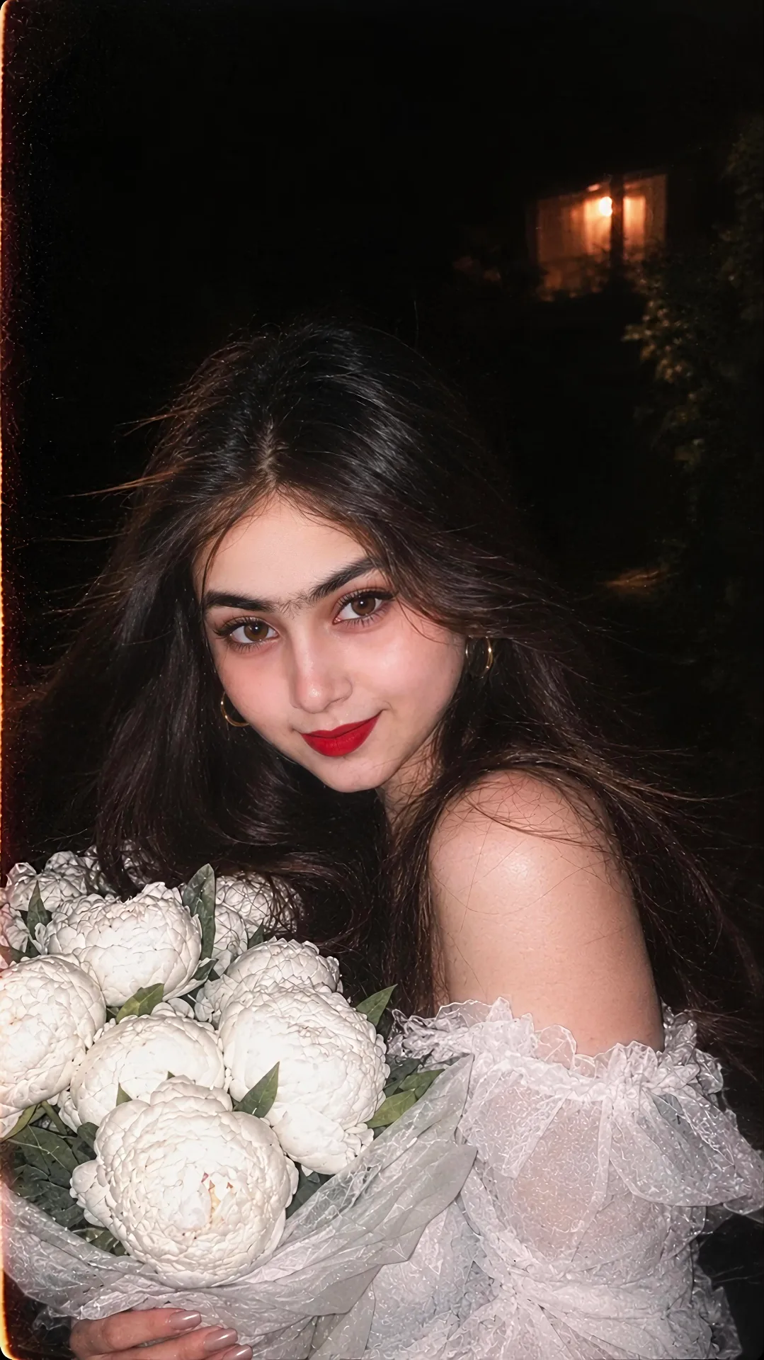 > A nighttime flash photography portrait of a beautiful young woman holding a large bouquet of white peonies wrapped in sheer paper.