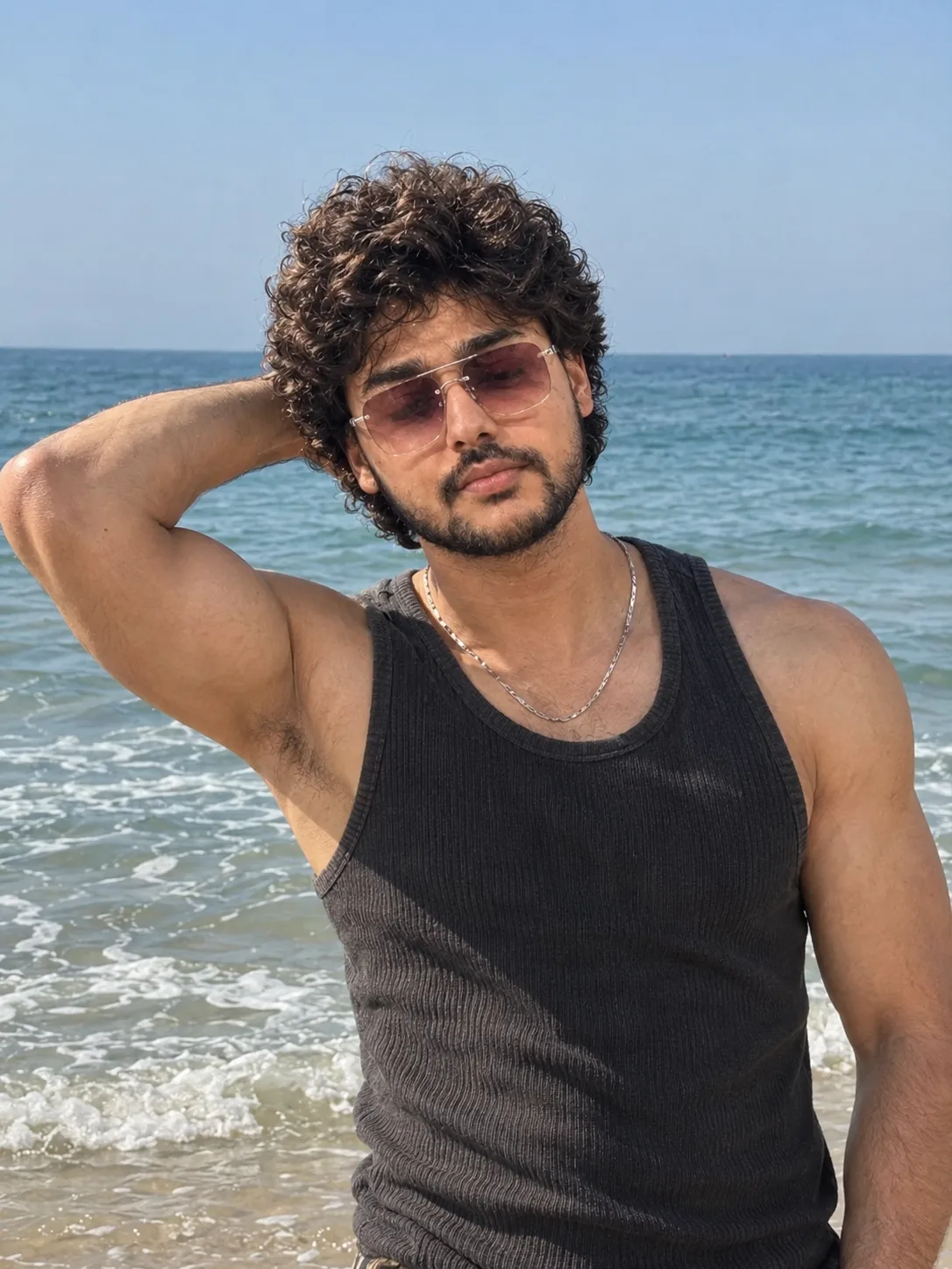 A stylish beach portrait of a young man with thick, curly, voluminous black hair and a neatly trimmed beard and mustache.