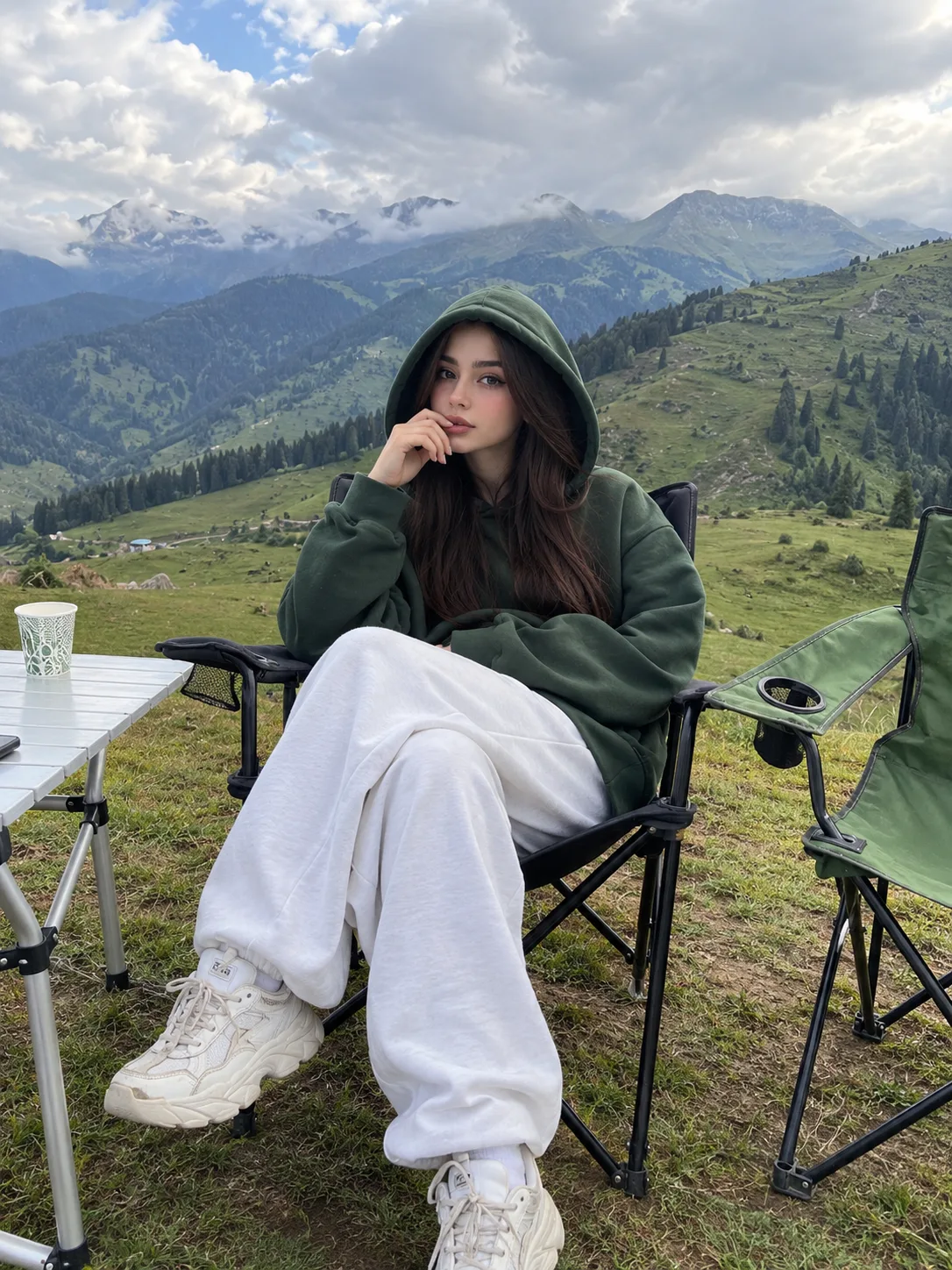 Candid camping photo in green mountains, beautiful young woman with long straight dark brown hair, sitting on black folding camping chair,
