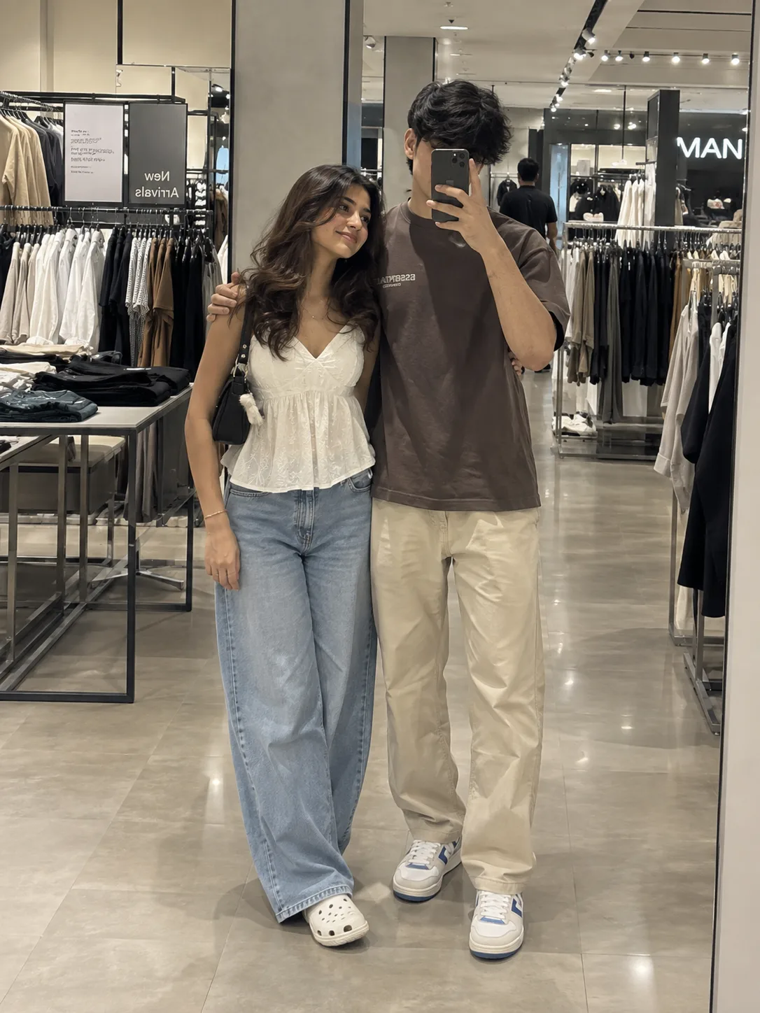 A realistic, full-body mirror selfie of a stylish young Indian couple inside a modern, well-lit clothing retail sto re.