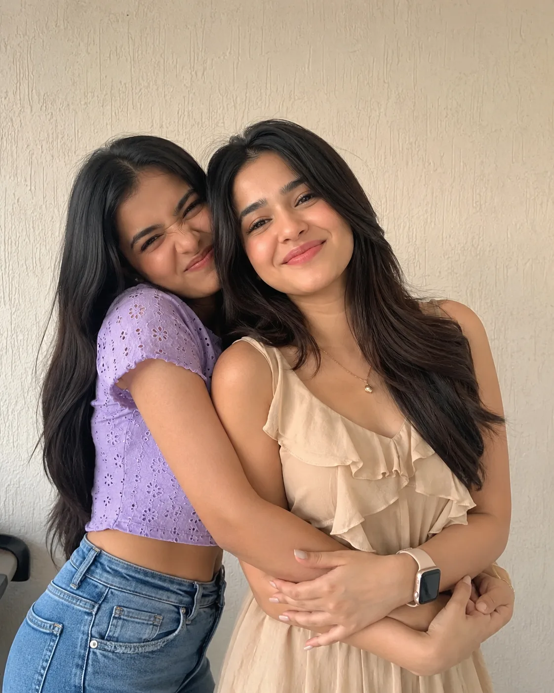 A candid, heartwarming portrait of two best friends posing playfully indoors against a clean cream-colored textured wall.