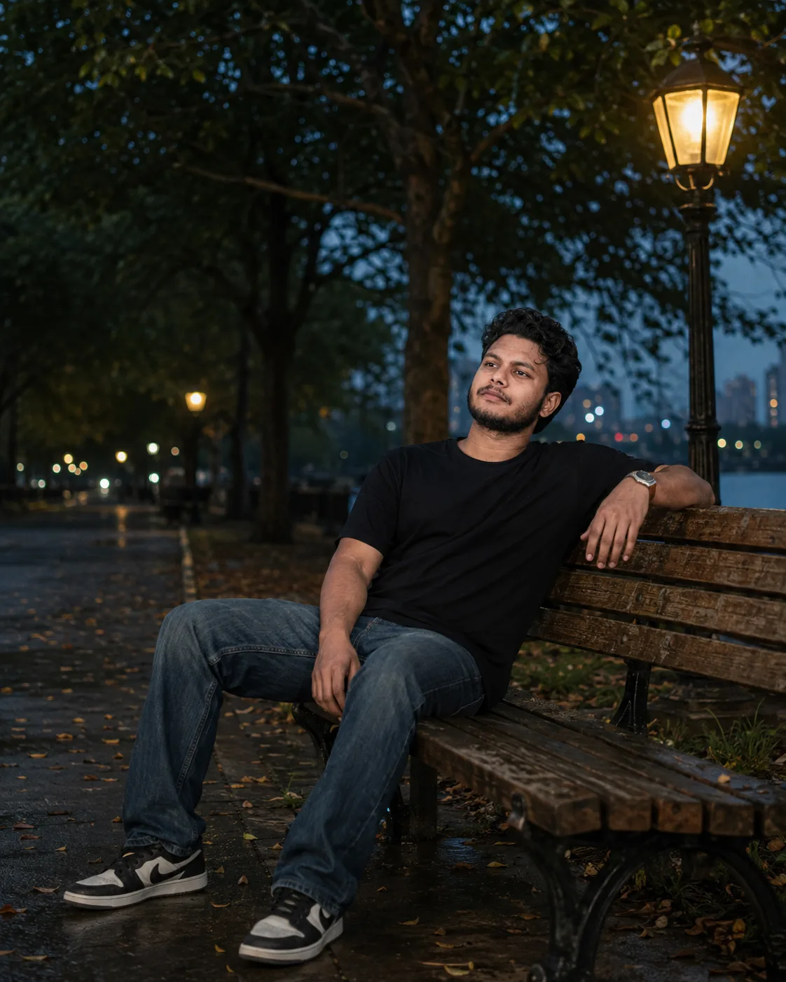 A cinematic night portrait of a young Indian man in his mid-20s with curly black hair, a short well-groomed beard and mustache, sitting