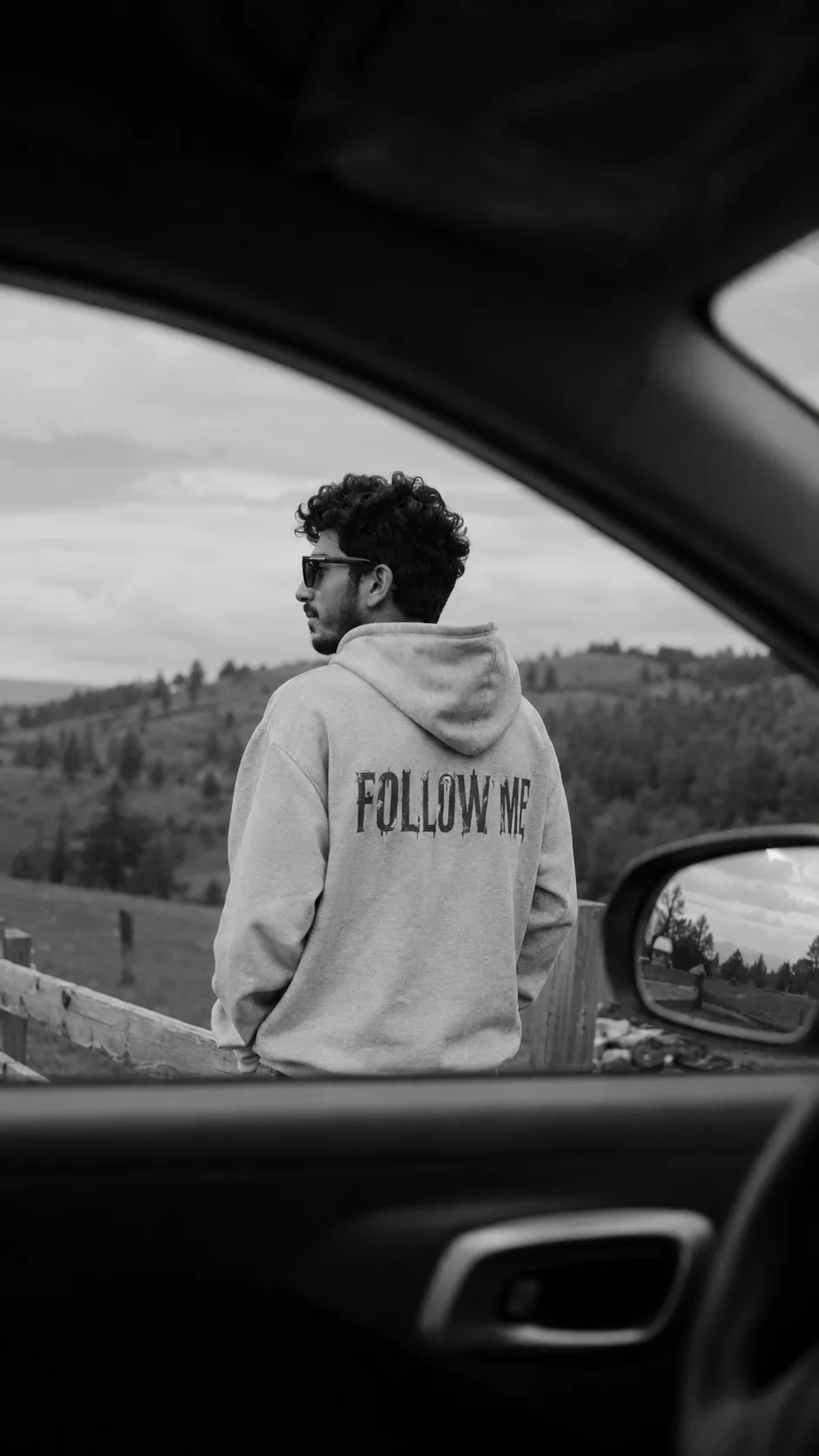 Ultra-realistic black and white cinematic photo of me standing outside near a wooden fence in an open rural area, viewed from inside a