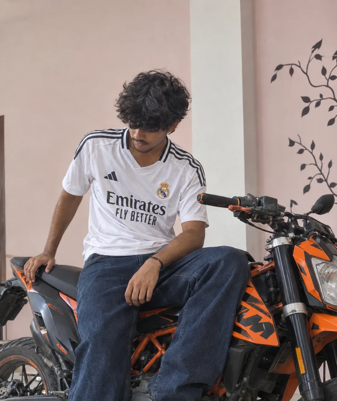 Ultra-realistic candid lifestyle photograph of a young South Asian man sitting casually on a sporty orange-and-black motorcycle indoors.