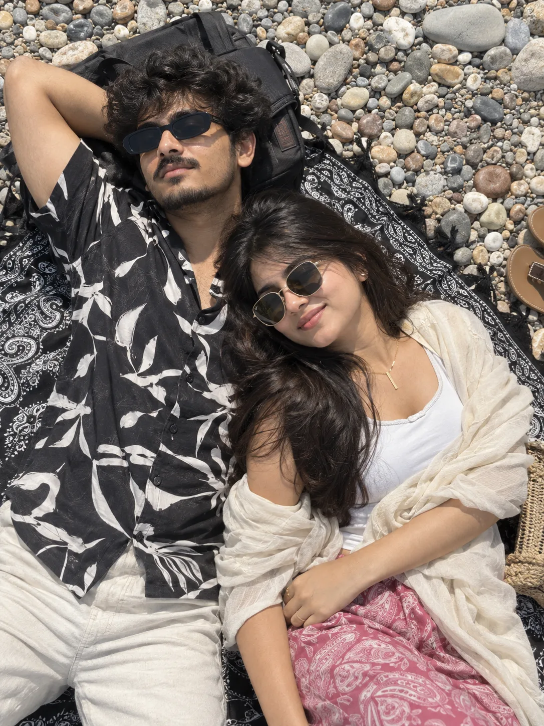 A hyper-realistic, top-down high-angle photograph of a relaxed young So uth Indian couple lying down together on a textured pebble beach