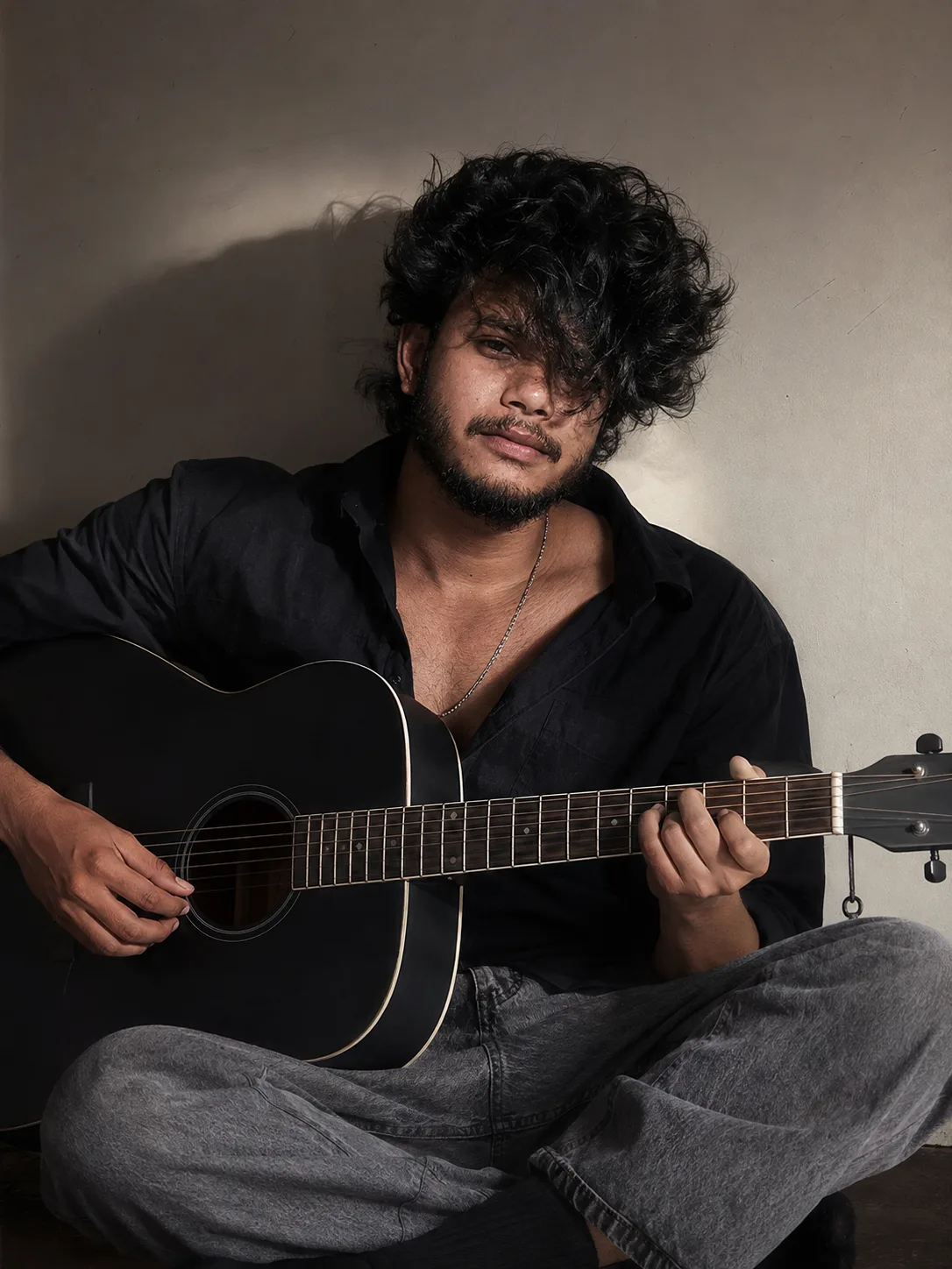 PROMPT A cinematic, moody portrait of a young man with thick, messy black fluffy hair covering part of his face, sitting cross-legged