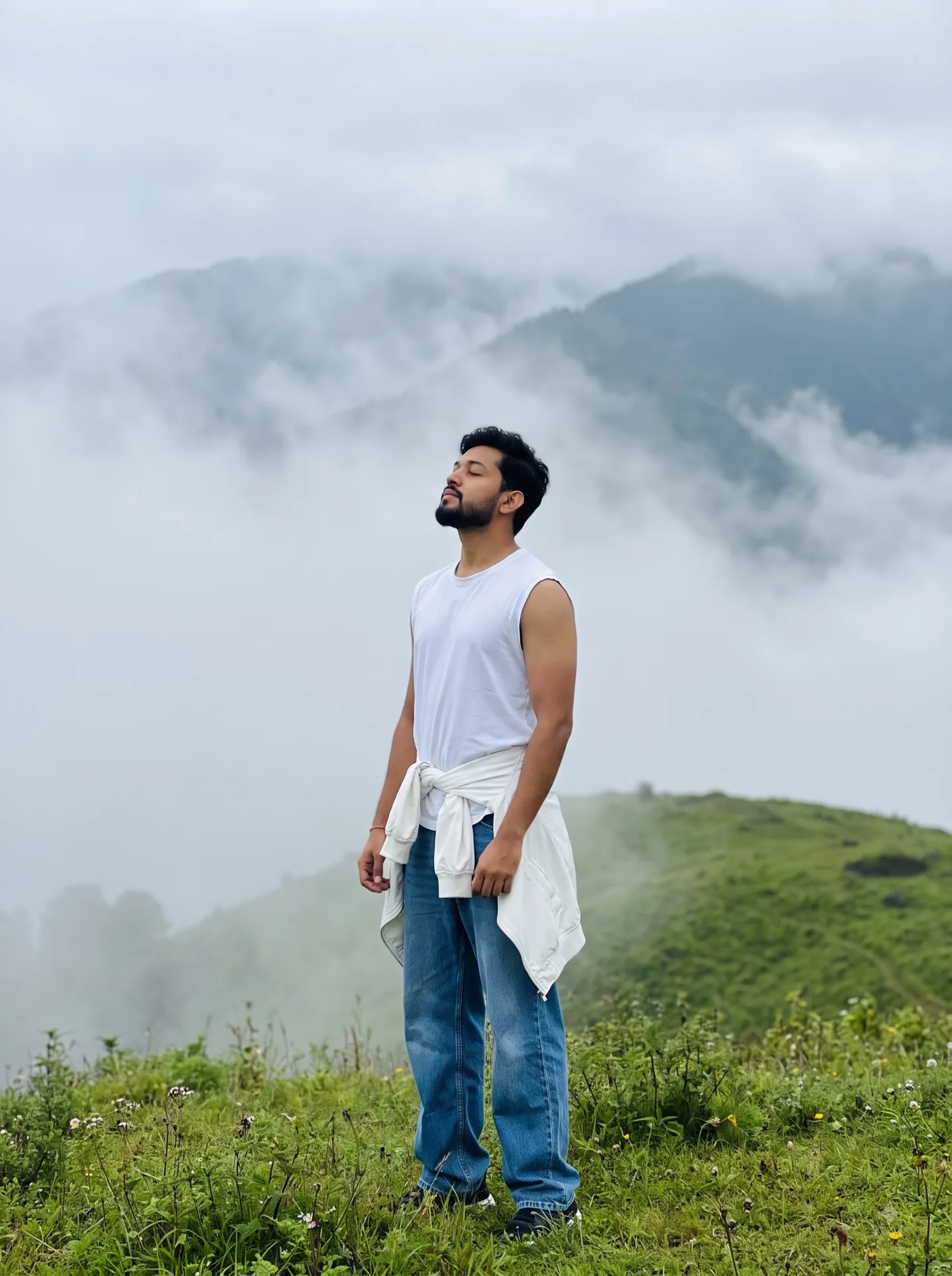 A STYLISH YOUNG MAN STANDS PEACEFULLY ON A LUSH GREEN HILLTOP SURROUNDED BY THICK FOG AND ROLLING CLOUDS HE WEARS CASUAL, TRENDY CLOTHES