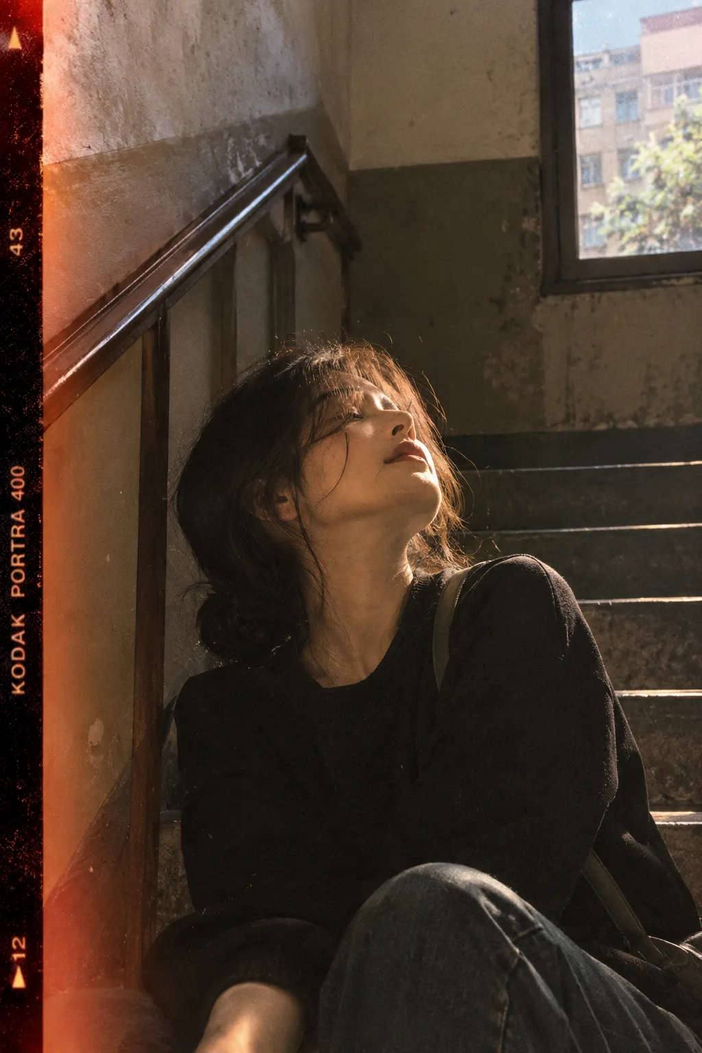 Flawed beauty, half-frame film texture, head tilted back catching the light, at the corner of a stairwell in an old residential building,