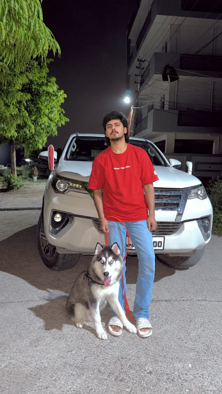 A cinematic nighttime street portrait of a young man standing confidently in front of a white luxury SUV, holding the leash of a Siberian