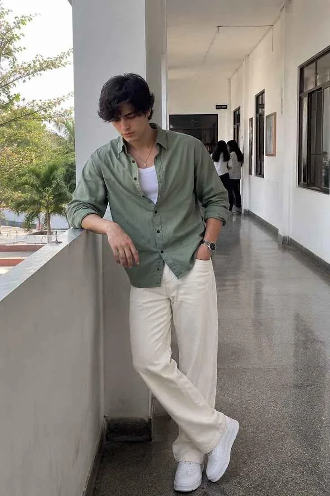 A high-quality full-body shot of a handsome young man with messy dark hair leaning against a balcony wall in a bright white corridor.