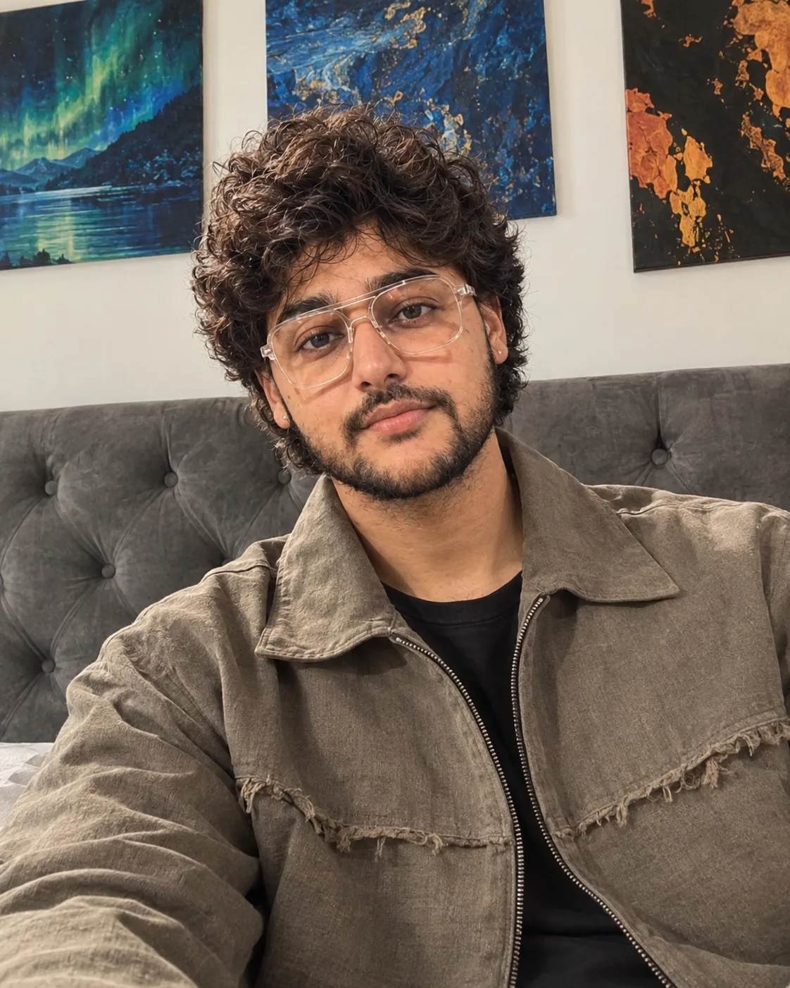 "A casual selfie-style portrait of a young man with thick, messy, tousled dark curly hair and a neatly trimmed black beard and mustache,