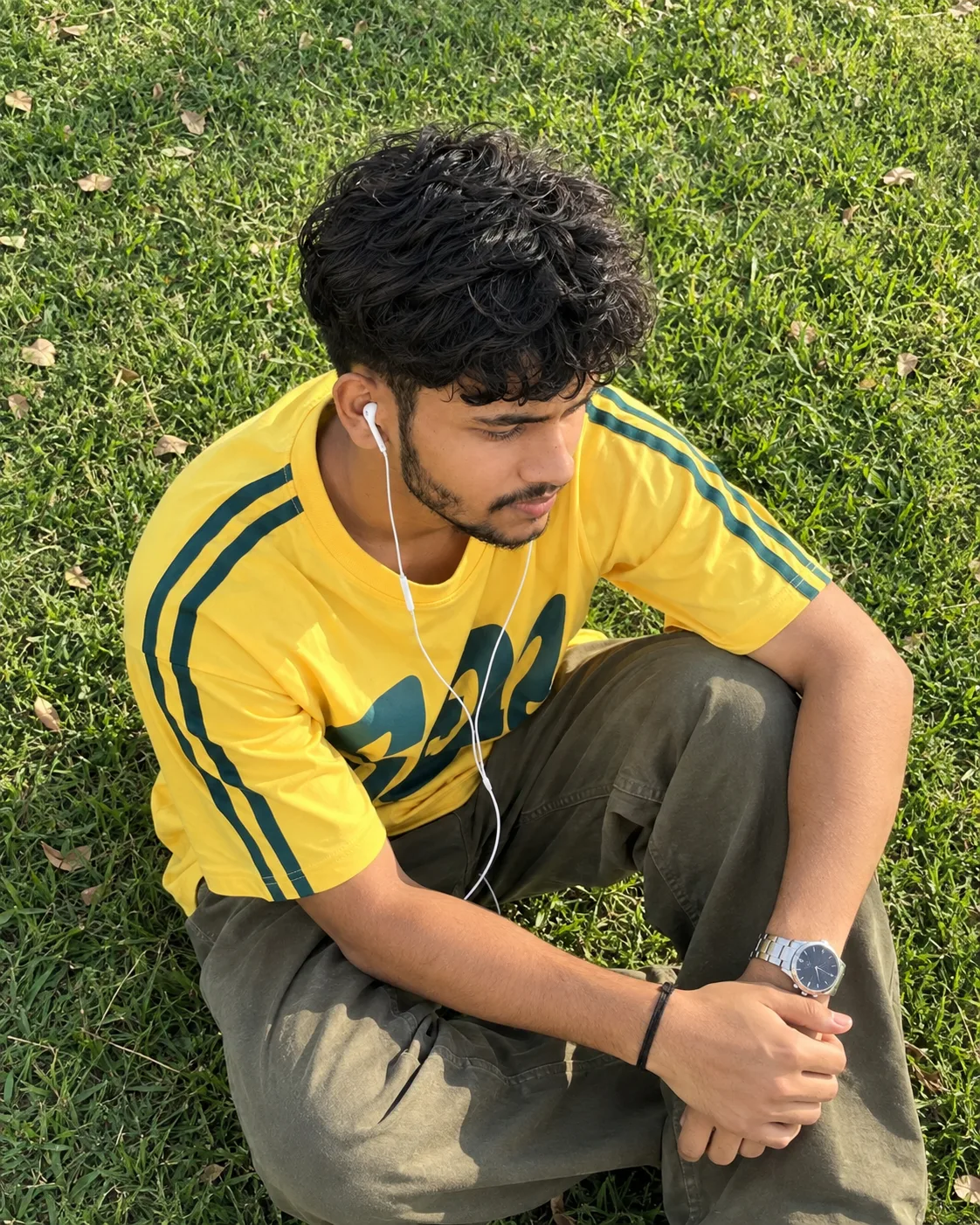 1. Ultra-realistic candid outdoor photograph of a young South Asian man sitting casually on a dense green grass lawn, captured from a high