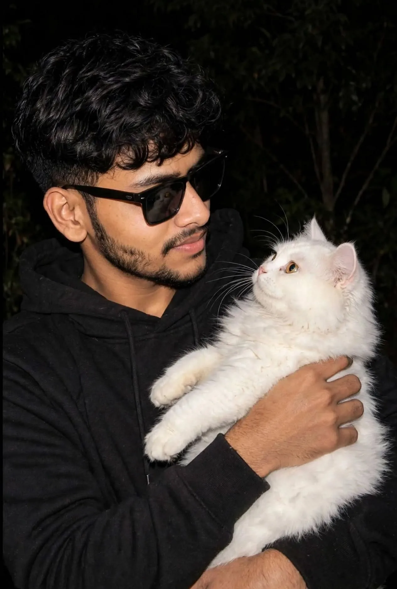 Candid night flash portrait of a young man with curly black hair, black sunglasses and black hoodie holding a fluffy white cat in his arms,
