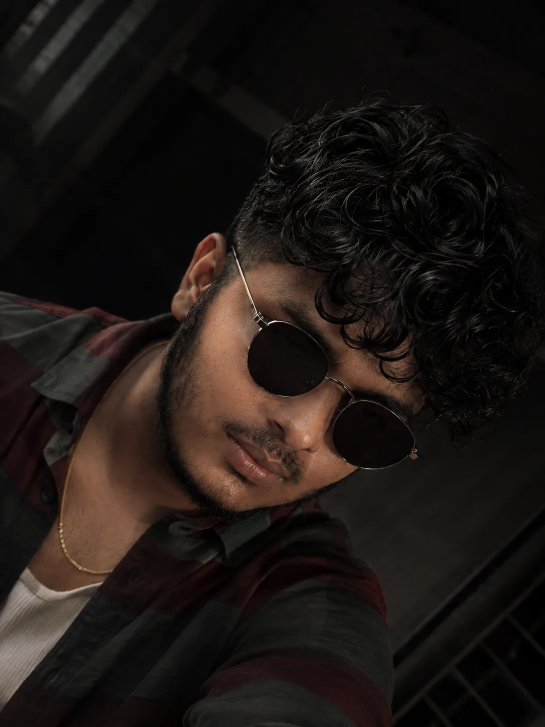 "A handsome South Asian man with a well-groomed beard, sporting large, messy, dark curly hair, and wearing round-framed, dark-lensed