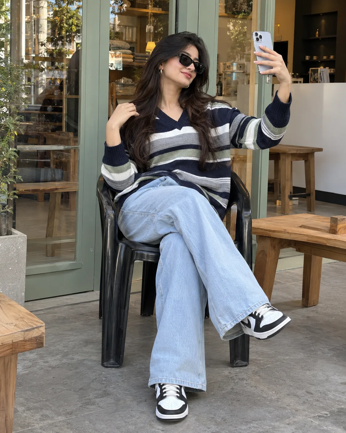 Ultra-photorealistic high-resolution DSLR photograph of a young Indian woman sitting casually on a black plastic chair outside a modern