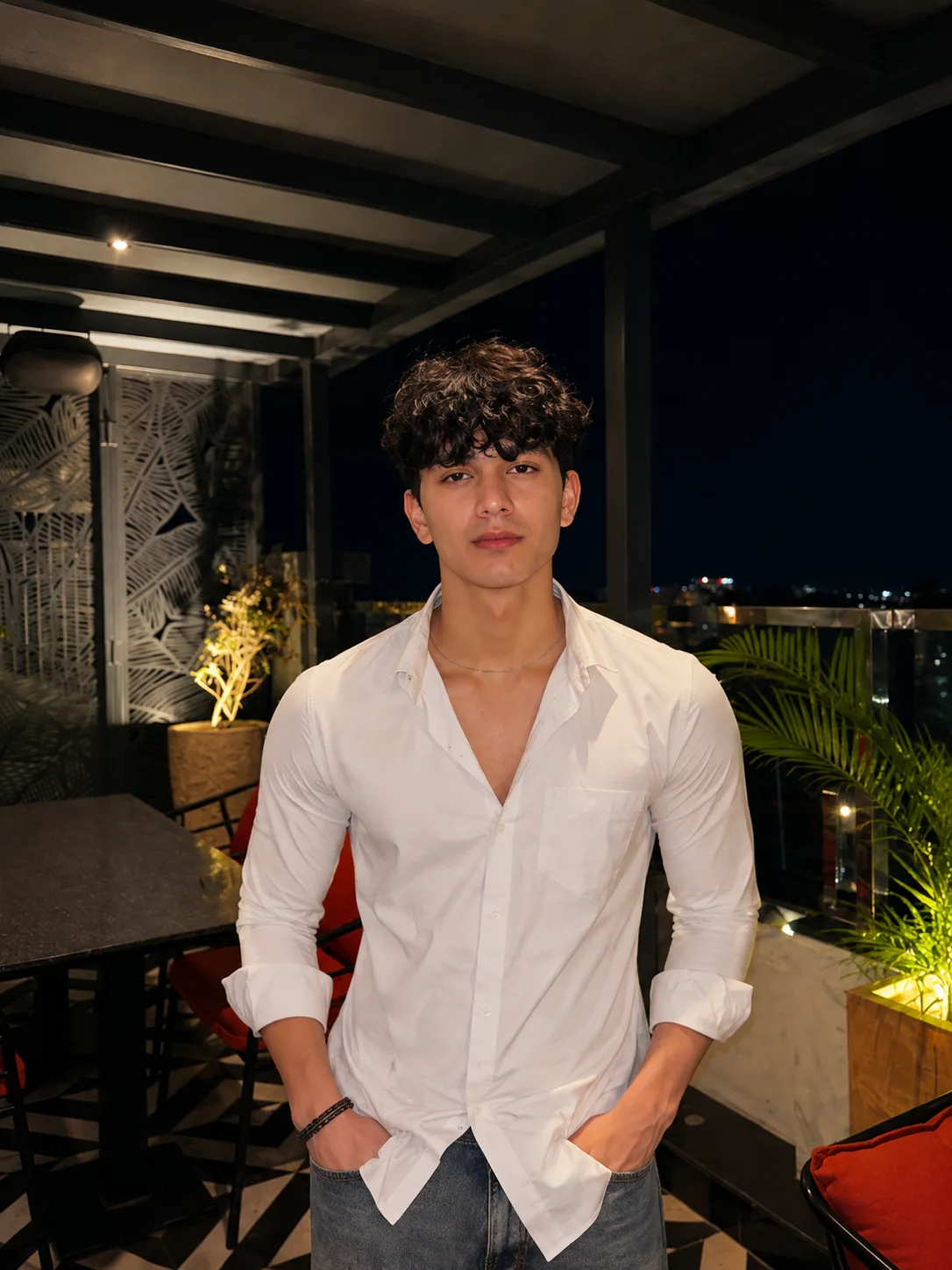 A strikingly handsome young male fashion model standing confidently on an exclusive luxury rooftop lounge during a warm metropolitan night.