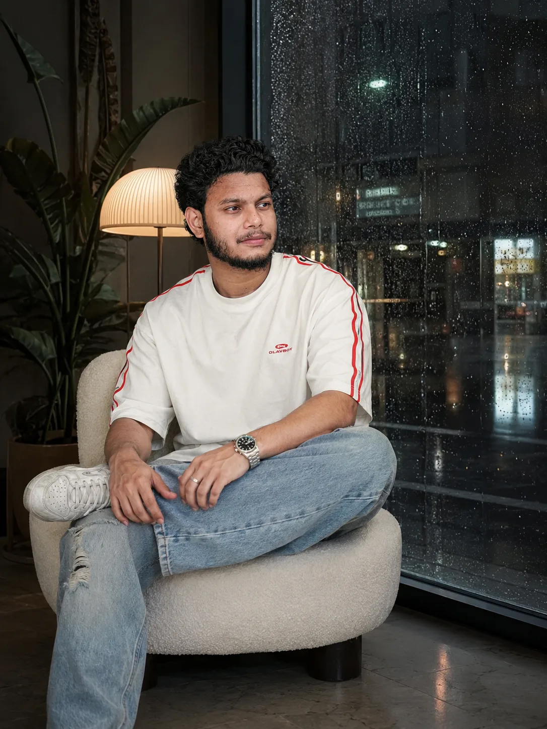 Prompt Barish Monsoon Stylish young Indian man sitting casually on a mini white boucle armchair,, versized white t-shirt with red stripe