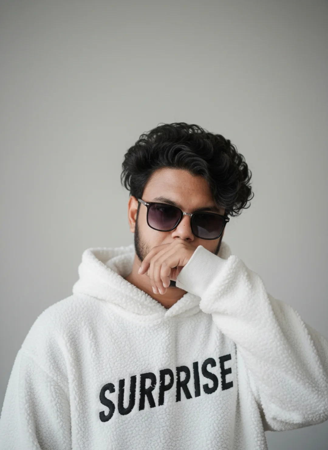 ** A young South Asian man with voluminous curly black hair and a short neat beard, wearing oversized black-framed gradient sunglasses,