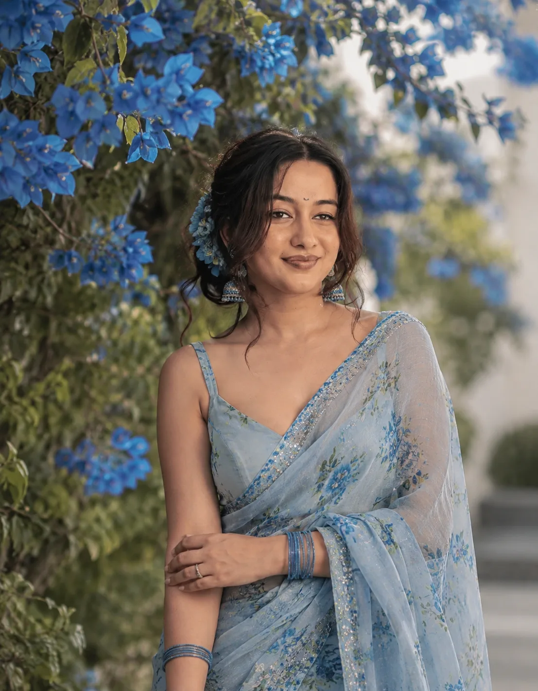Generate a hyper-realistic, cinematic portrait of the uploaded beautiful woman standing gracefully in a garden filled with vibrant blue