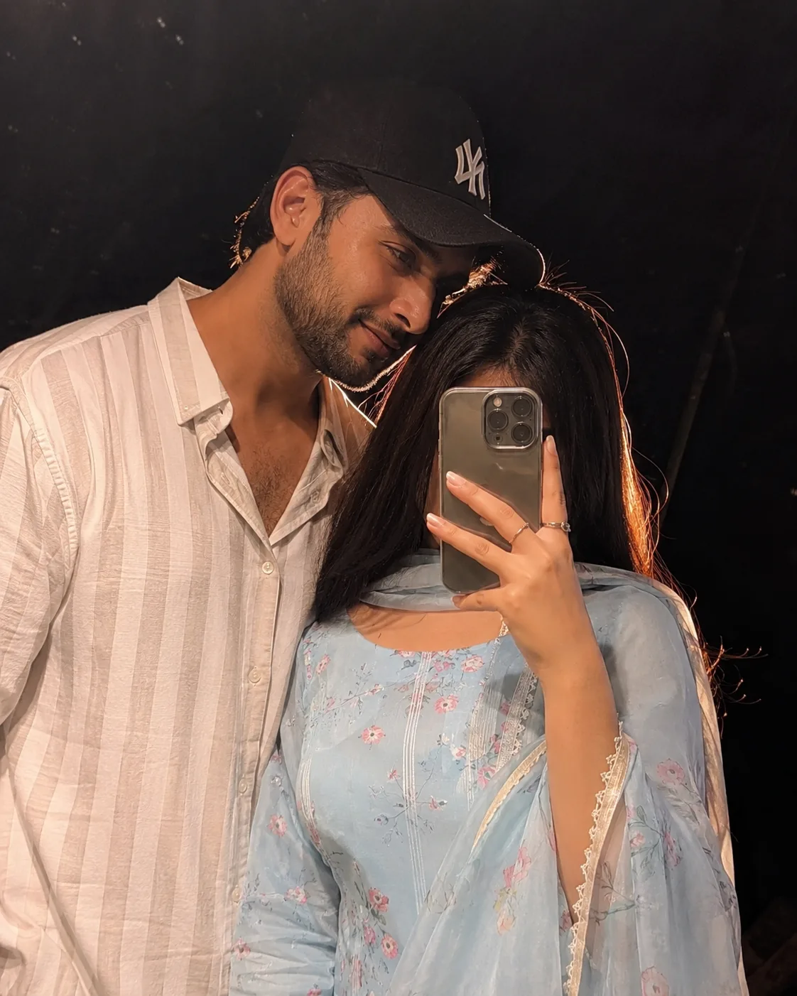 Aesthetic nighttime mirror selfie of a young couple, boy wearing a black NY cap and white striped casual shirt, trimmed beard, soft smile,