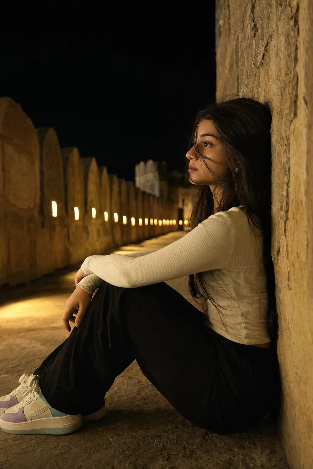 Ultra-realistic cinematic nighttime portrait of a young South Asian woman sitting alone against a textured golden-beige stone wall in a