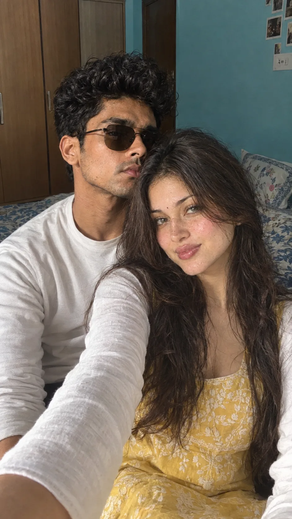 A natural, ultra-realistic indoor selfie of a young couple sitting closely together on a bed in a cozy bedroom.