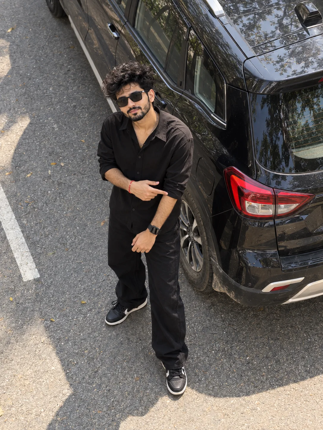A high-angle, full-body portrait of a smiling young man standing on a sunlit asphalt road next to the rear passenger side of a parked black