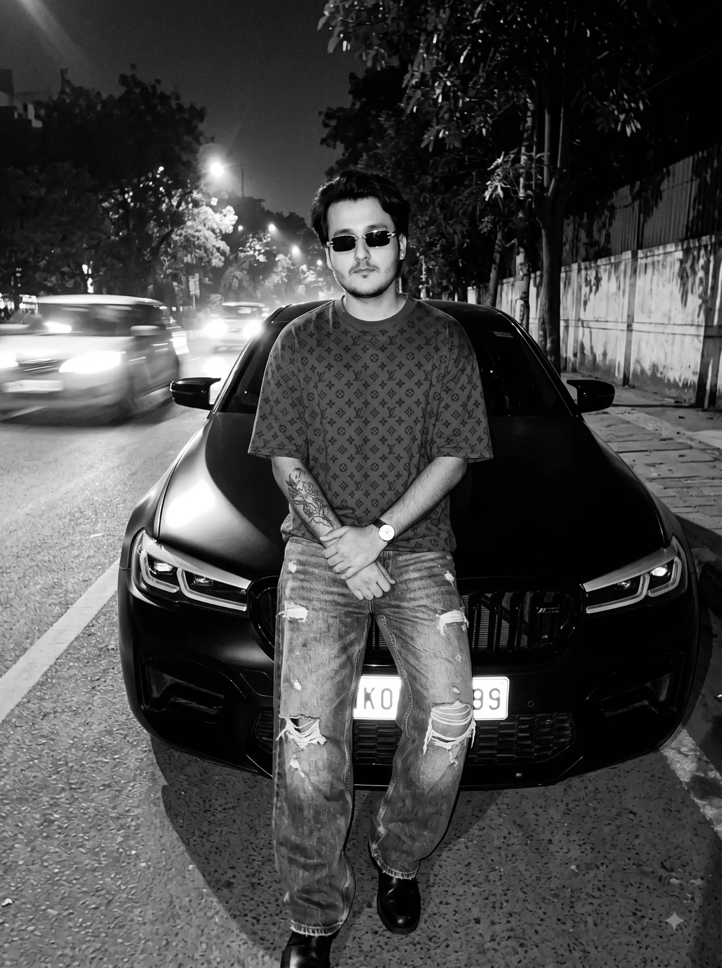 Black and white grainy flash photography at night, vertical full body shot of a young South Asian man in his mid-20s with stylish black