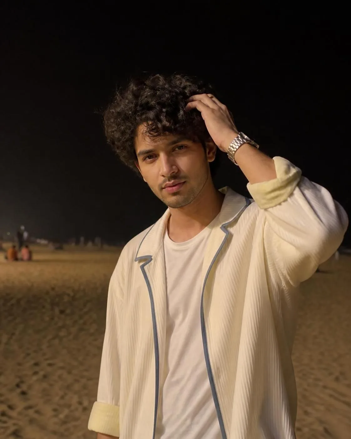 Create a realistic nighttime outdoor portrait of a young man standing on a sandy beach or open shoreline.