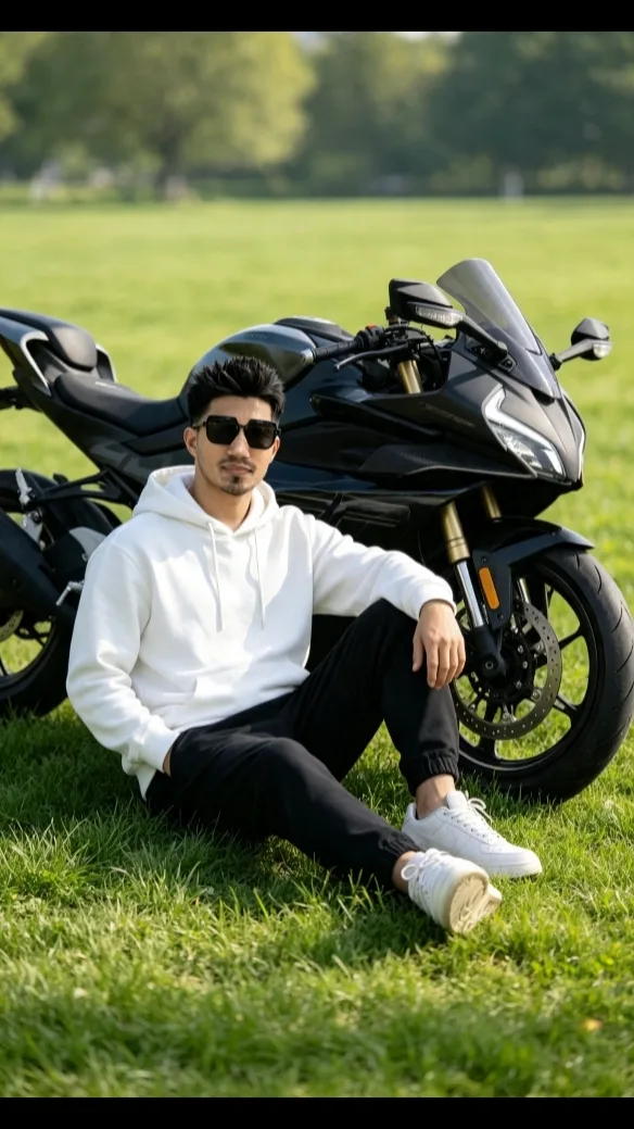 A stylish young man sitting casually in a lush green grassy field, wearing a white hoodie, black pants, and white sneakers, looking