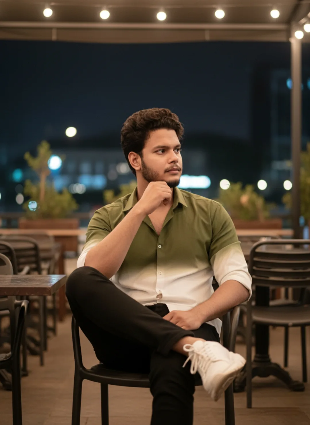 A young Indian man with curly dark hair and a short beard sits casually on a black outdoor chair at night, legs crossed, right hand resting