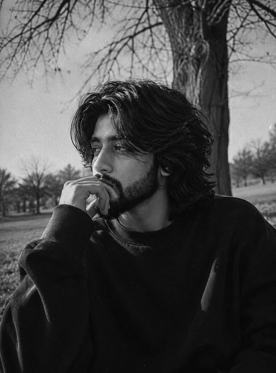 A high-contrast black and white vintage film portrait of a man outdoors sitting in a pensive, contemplative pose turned to a three-quarter