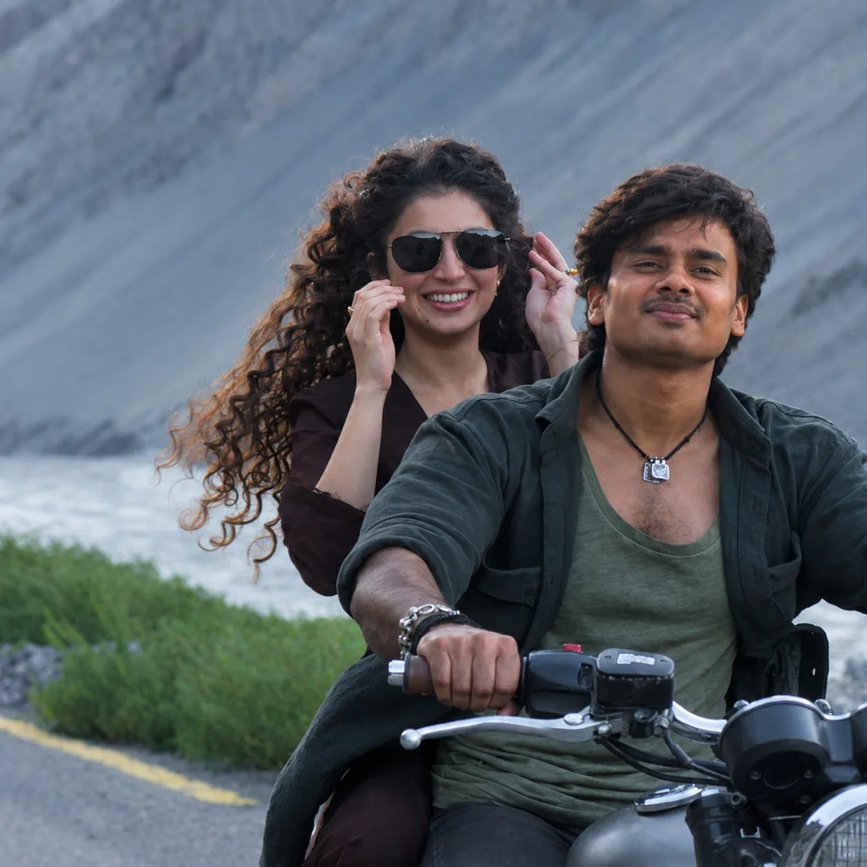 Create a cinematic romantic adventure poster featuring a young couple riding a classic motorcycle along a dramatic coastal mountain road.