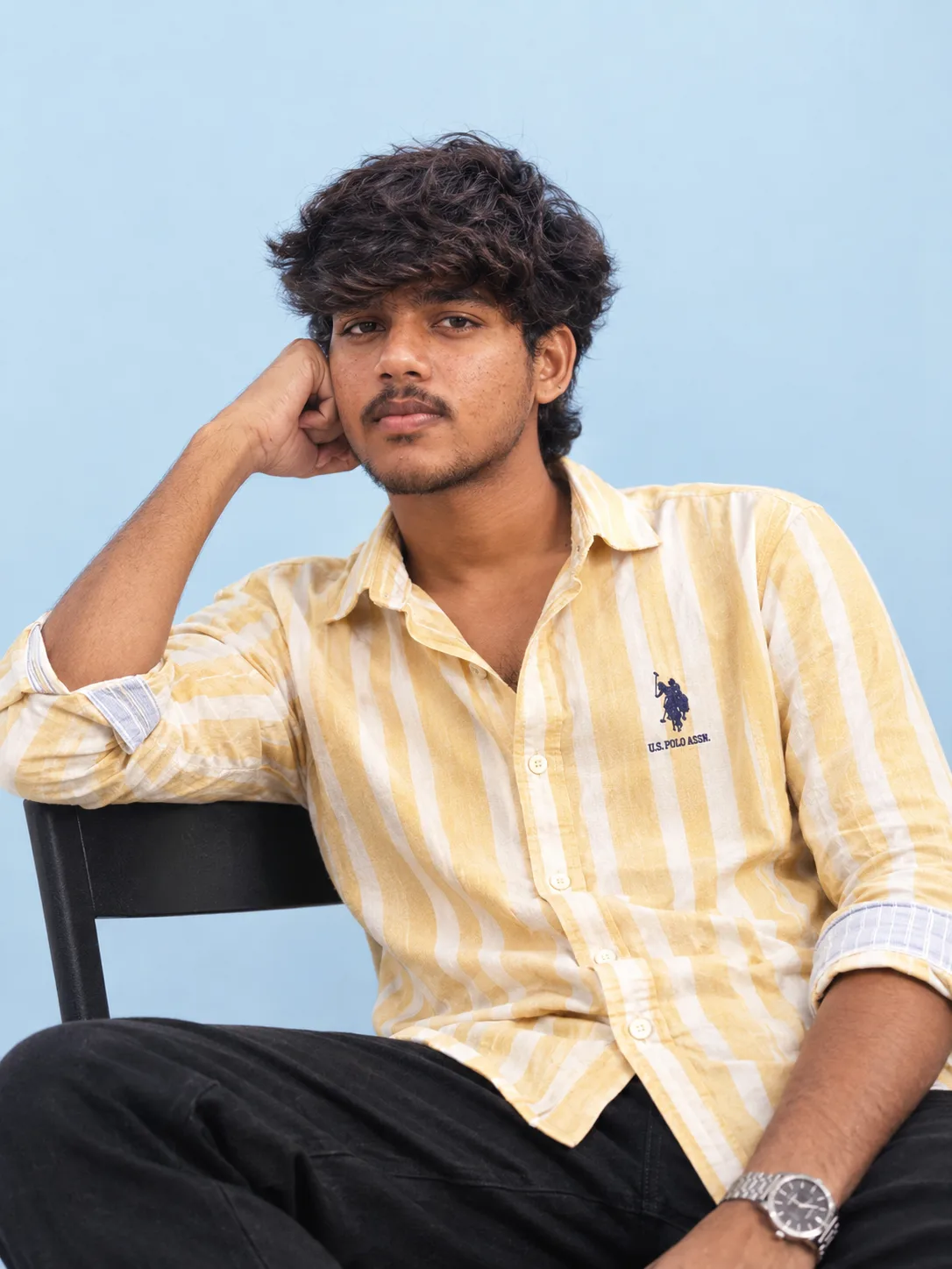 Ultra-realistic cinematic fashion portrait of a stylish young South Asian man sitting casually on a simple black chair against a clean