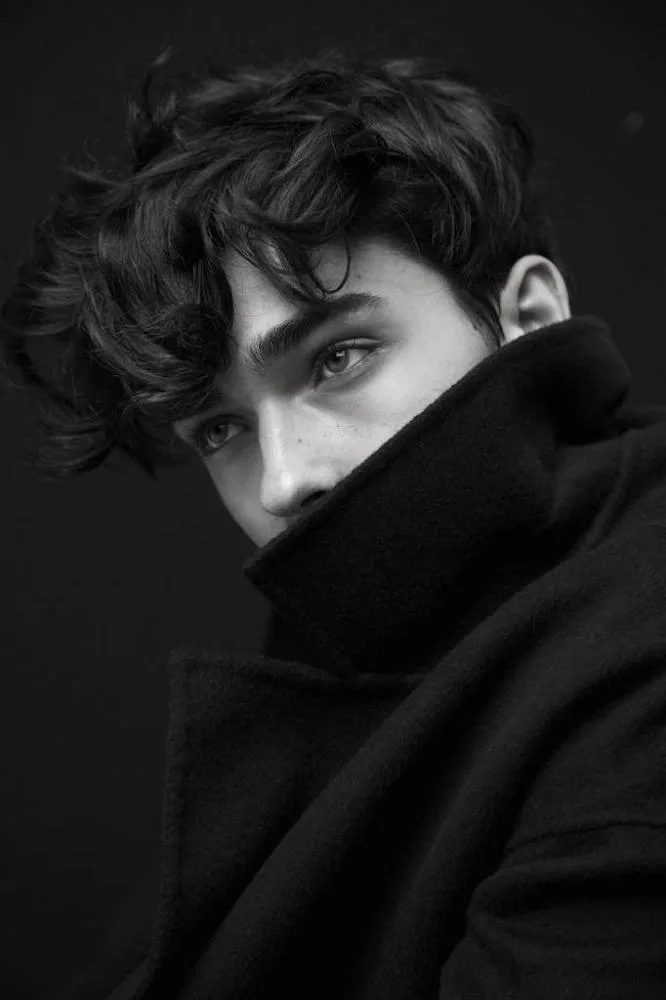 A striking black and white close-up studio portrait of a handsome young man with messy, wavy dark hair softly falling over his forehead.