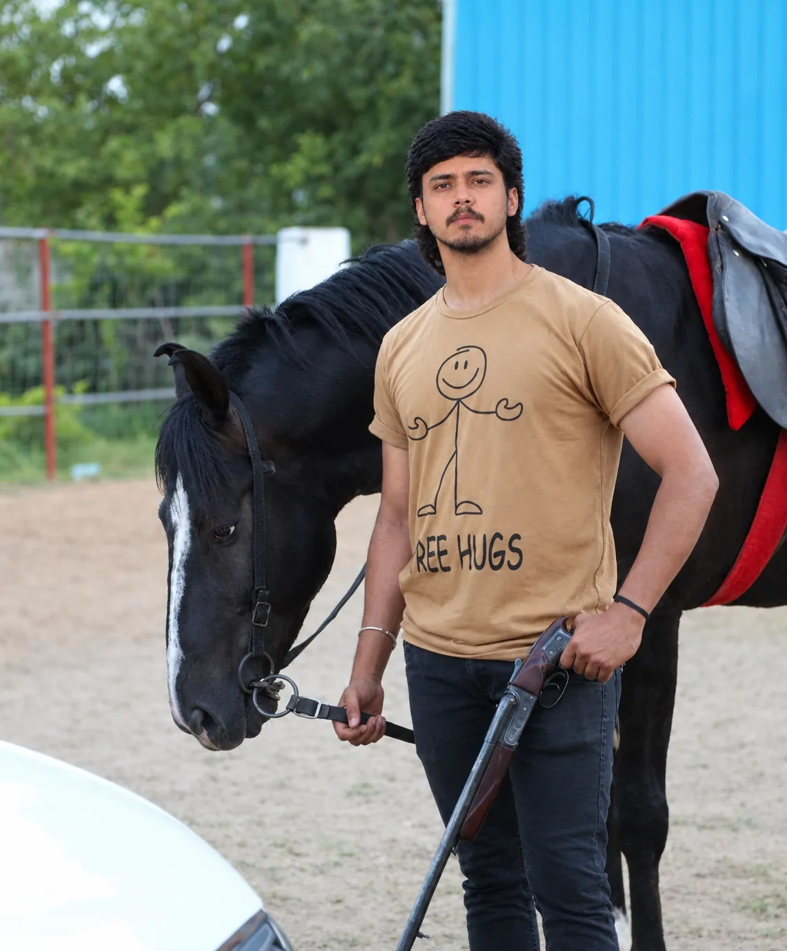 Photorealistic outdoor portrait of a young South Asian man standing beside a large black horse in an equestrian paddock.