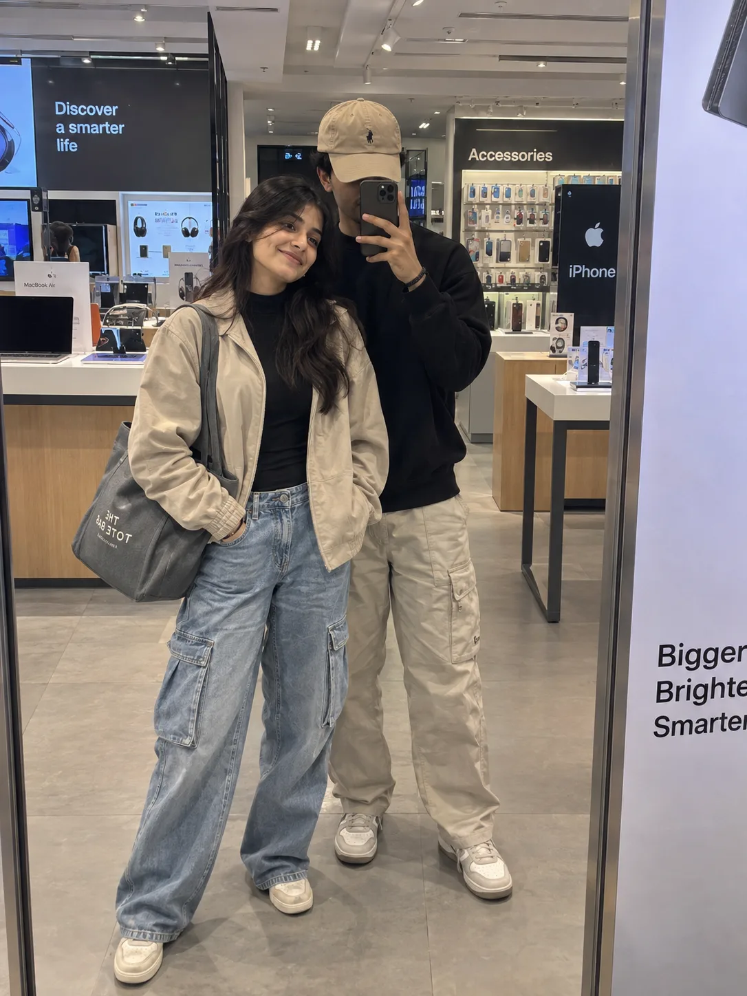 A realistic, candid full-body mirror s elfie of a cheerful young Indian couple posing inside a modern, brightly....