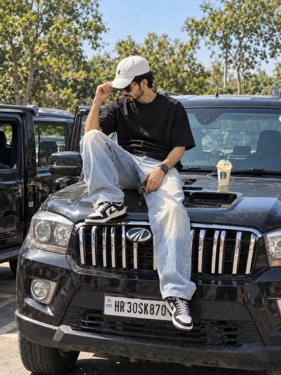 Prompt👇🏻 Photorealistic photo of a young Indian man in his mid-20s sitting casually on the hood of a black Mahindra Scorpio SUV, front