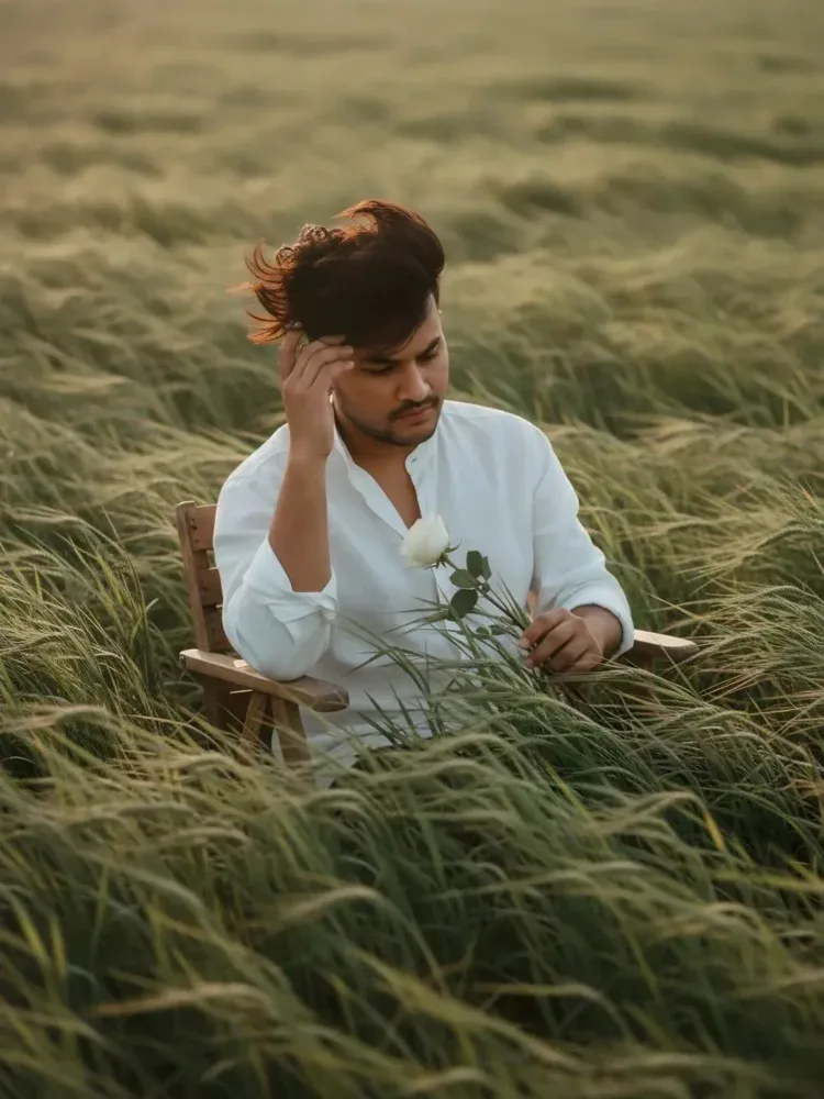 Use the attached photo to create a cinematic editorial portrait of a man sitting on a simple chair in vast, wind-swept green grass fields.