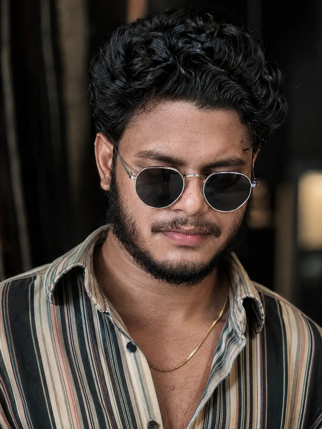 A close-up portrait of a young South Asian man with medium-dark skin tone, looking slightly downward with a calm, confident expression.