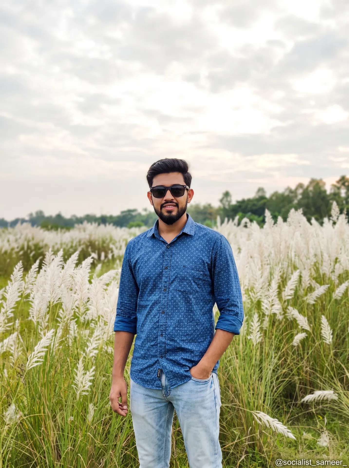 Realistic wide shot of a young South Asian man standing confidently in a lush Kashphool field under a bright cloudy autumn sky.