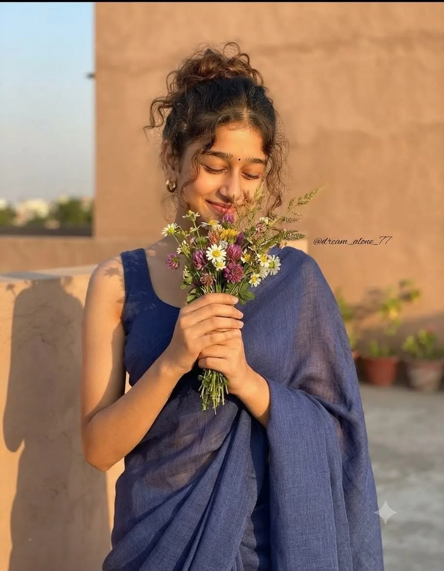 A gorgeous South Asian woman wearing a modern indigo blue linen saree with a matching sleeveless blouse, standing on a sunlit rooftop.